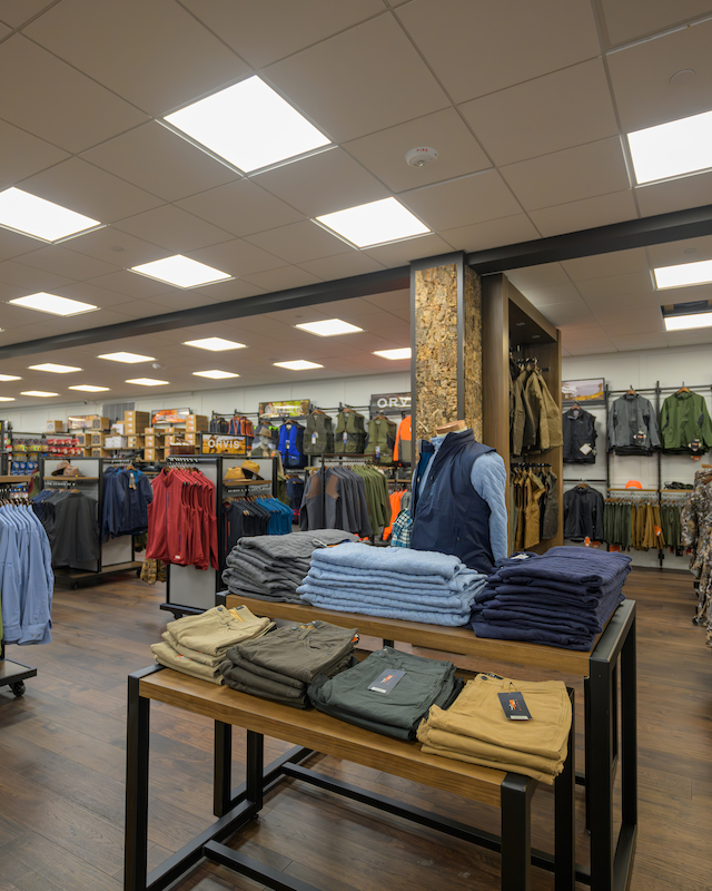 The image shows a retail store interior with clothing racks, neatly folded shirts, and outdoor gear displayed under bright lighting.