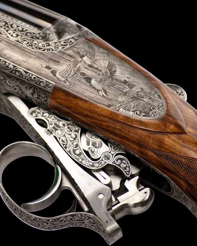 The image shows a close-up of a beautifully engraved firearm with intricate metalwork and wooden details, likely a shotgun.