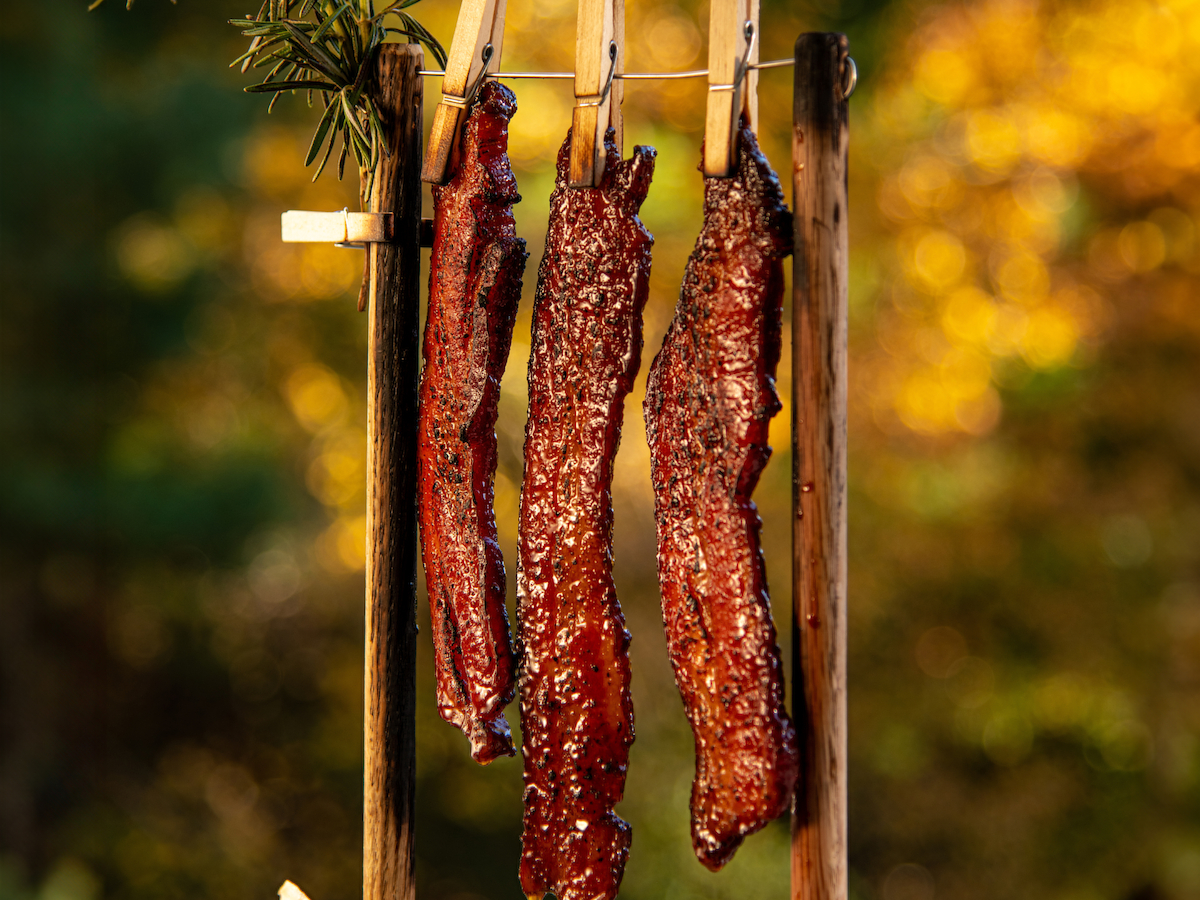 Three strips of bacon hanging on clothespins with a pickle and lemon wedge on a wooden plate stand against a blurred autumn background.