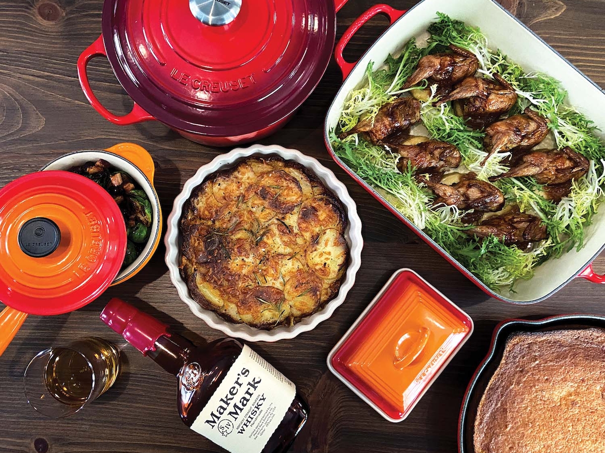 The image shows a table with various dishes including a pie, roasted vegetables, and a loaf of bread, along with a bottle of Maker's Mark.