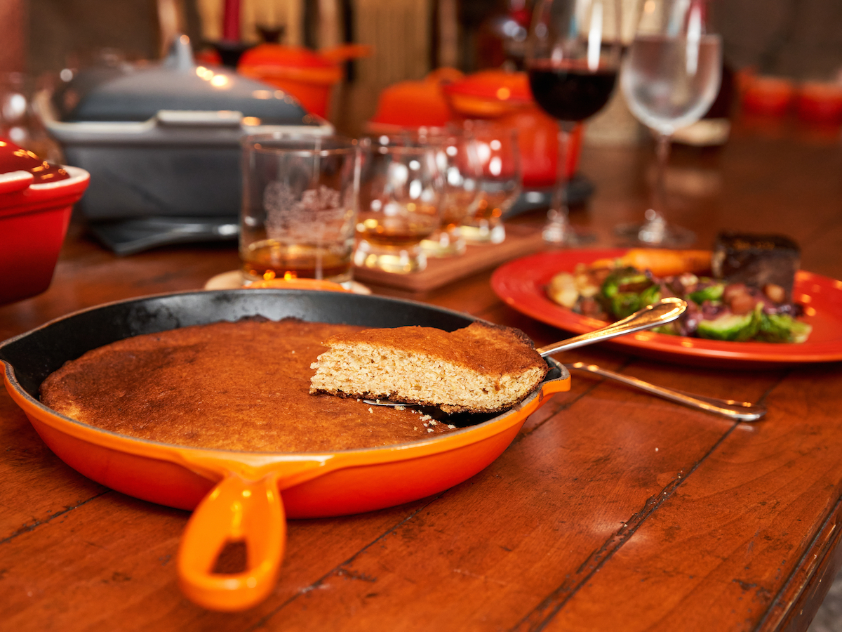 A table with food and drinks, featuring cornbread in a skillet, a red plate with a meal, and various glasses, including wine, in the background.