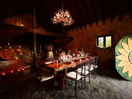 A rustic dining room with a wood table, chairs, antler chandelier, stone walls, and a sunflower decoration. Cozy and inviting ambiance.