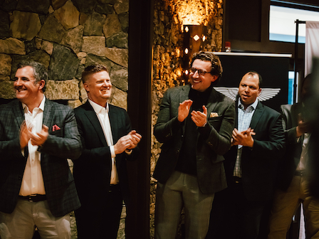 A group of four people are standing and applauding in a warmly lit room with a stone wall background.