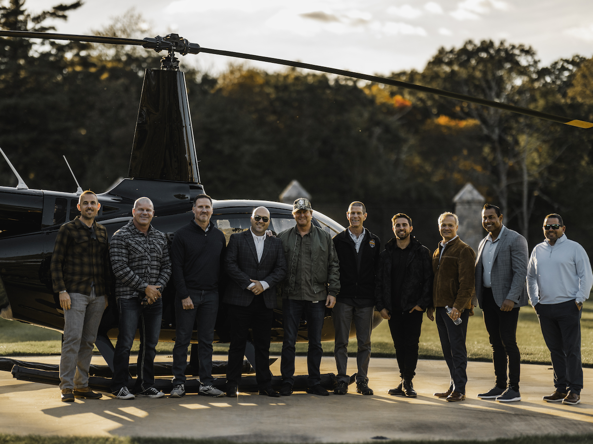 A group of people standing in front of a helicopter on a sunny day, with trees in the background.