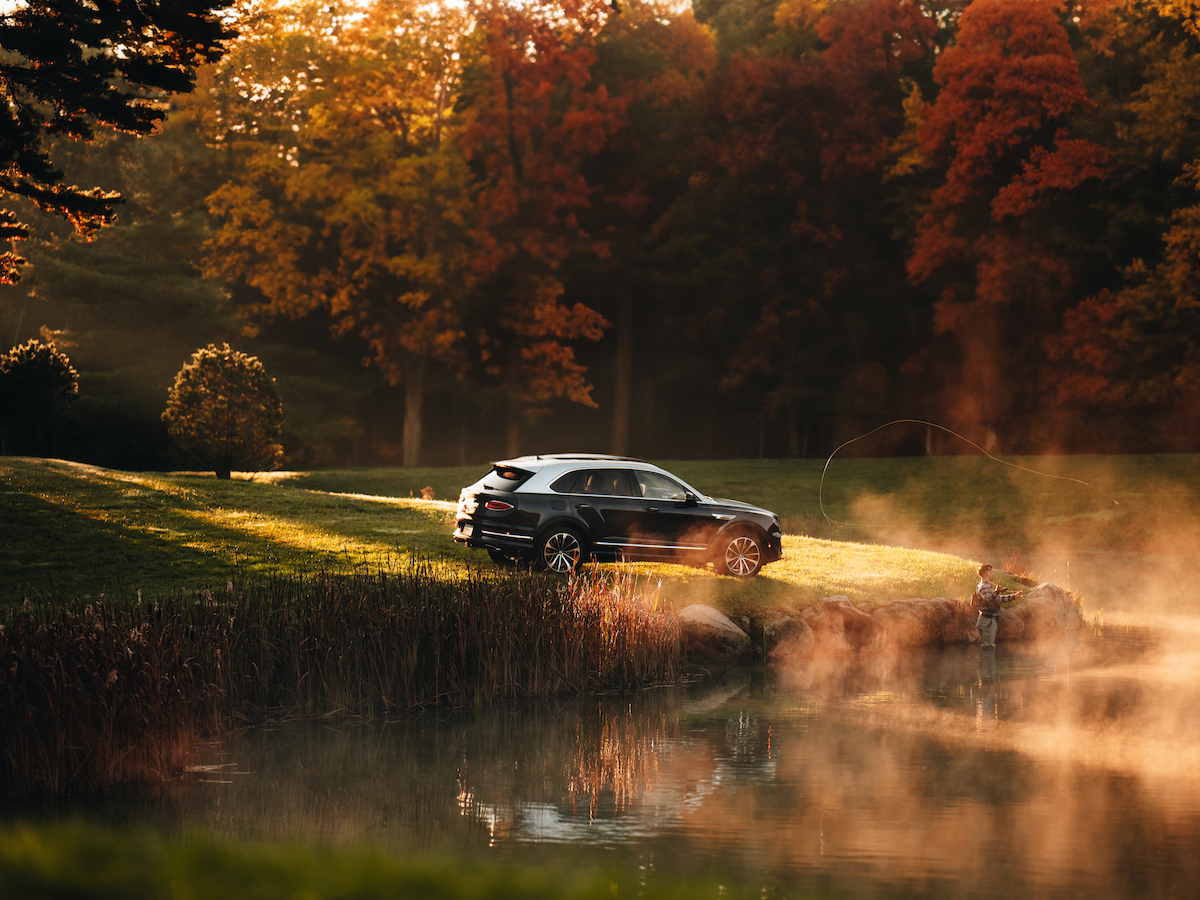 A car is parked near a misty pond in a park, surrounded by autumn trees with vibrant foliage, under warm sunlight.